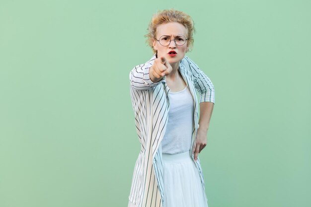 Eh, tú. Retrato de una joven rubia seria con camisa blanca, falda y blusa a rayas y anteojos de pie, mirando y apuntando a la cámara. tiro de estudio interior aislado sobre fondo verde claro