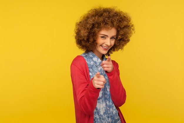 Eh, tú. Retrato de una mujer feliz y alegre con el pelo rizado y esponjoso con ropa informal apuntando a la cámara, notando e indicando el dedo, eligiendo la afortunada. tiro del estudio aislado sobre fondo amarillo