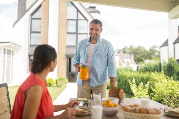 Ehemann bringt Saft. Fürsorglicher lächelnder Ehemann, der mit seiner Frau draußen etwas Orangensaft zum Frühstück bringt?