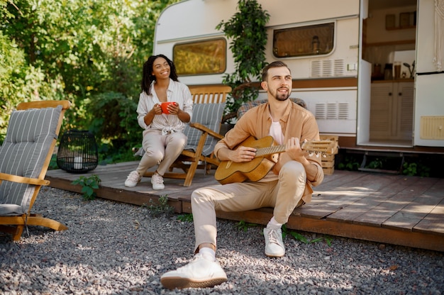 Ehemann mit Gitarre in der Nähe von Wohnmobil, Abenteuer auf Rädern, Camping in einem Anhänger. Mann und Frau reisen mit dem Van, Urlaub mit dem Wohnmobil, Wohnmobile in Wohnmobilen