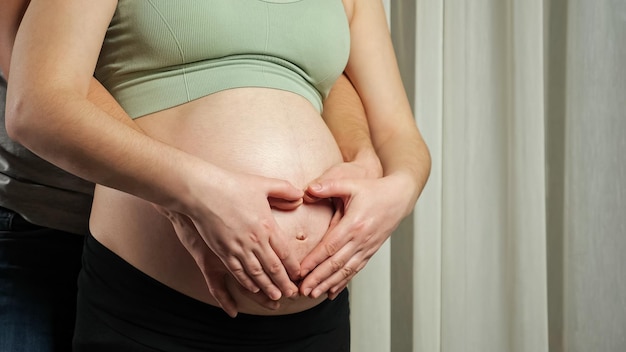 Ehemann und Ehefrau machen ein Herzzeichen, das den schwangeren Bauch umarmt