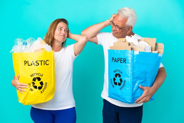 Ehepaar mittleren Alters, das eine Recyclingtüte voller Papier und Kunststoff hält, die auf weißem Hintergrund isoliert ist und beim Kopfkratzen Zweifel hat