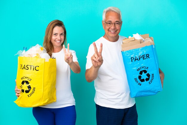 Ehepaar mittleren Alters, das eine Recyclingtüte voller Papier und Kunststoff hält, isoliert auf weißem Hintergrund, lächelt und zeigt Victory-Zeichen