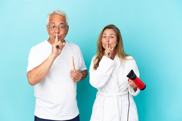 Ehepaar mittleren Alters, das Trockner und Zahnbürste einzeln auf blauem Hintergrund hält und ein Zeichen der Stille zeigt, die den Finger in den Mund steckt