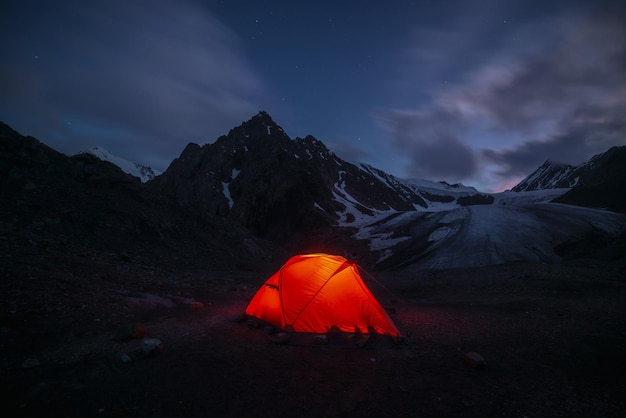 Ehrfürchtige Berglandschaft mit lebhaftem orangefarbenem Zelt in der Nähe einer großen Gletscherzunge unter Wolken im nächtlichen Sternenhimmel Zelt leuchtet durch orangefarbenes Licht mit Blick auf Gletscher- und Bergsilhouetten in sternenklarer Nacht