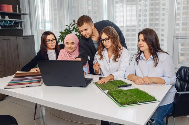 Ehrgeizige aktive junge Gruppe der besten Arbeiter, die an einem weißen Tisch sitzen und Projekte analysieren und planen. Attraktives Mädchen in einem rosa Hijab im Büro. Konzept für harte Arbeit