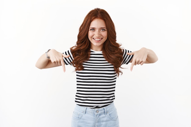 Foto ehrgeiziges gutaussehendes lächelndes glückliches mädchen mit roten haaren in gestreiftem t-shirt, das nach unten zeigt, gute werbung zeigt, amüsiert grinst, werbung einführt, weiße wand