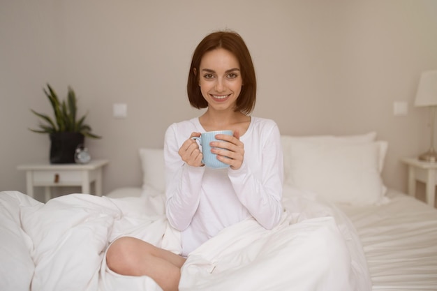 Ehrliches Foto einer fröhlichen jungen Frau, die im Bett Kaffee trinkt
