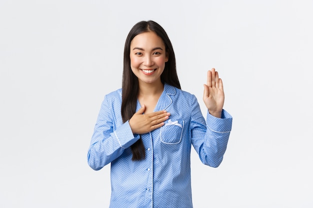 Ehrliches und aufrichtiges schönes asiatisches Mädchen im blauen Pyjama, das eine Hand hebt und Handfläche auf Herz hält, um Versprechen zu machen, Wahrheit zu sagen oder Eid zu geben, lächelt, als würde es Marmeladen über weißer Wand tragen