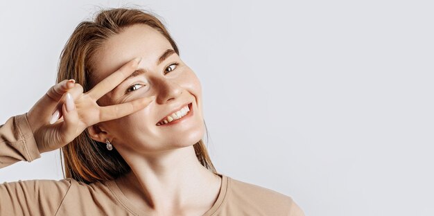 Ei sorria você. Linda jovem sorri e mostra o gesto de vitória da paz como brincar em um fundo branco e isolado. Uma mulher aponta para uma ideia, um lugar para anunciar. Morena fofa positiva