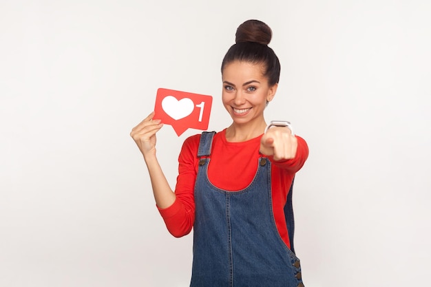 Ei você, curta e acompanhe conteúdos interessantes. Retrato de menina elegante positiva com coque de cabelo em macacão jeans segurando o ícone de coração de mídia social e apontando para a câmera. tiro de estúdio interno, isolado