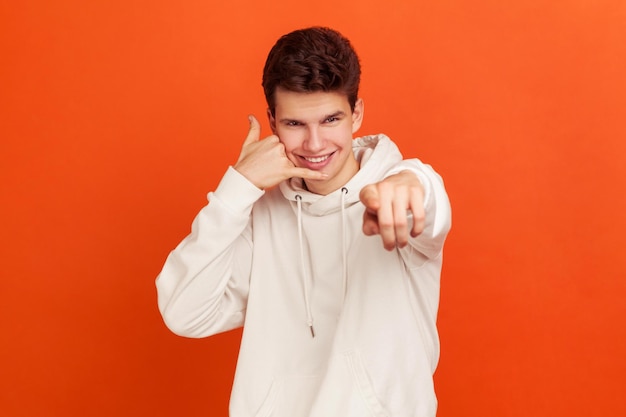 Ei você, me ligue. Adolescente brincalhão bonito com capuz estilo casual e com penteado elegante flertando mostrando gesto de chamada. Tiro de estúdio interior isolado em fundo laranja