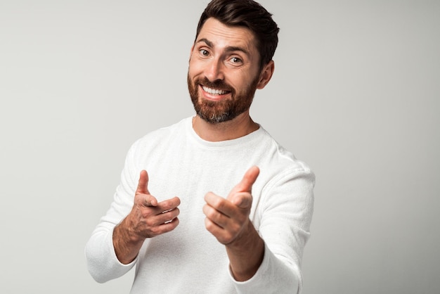 Foto ei, você retrato de homem barbudo feliz na camisa branca, apontando com os dedos para alguém contra um fundo cinza claro foto de estúdio