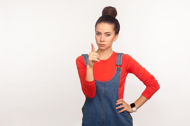 Ei você. Retrato de menina zangada mandona com coque de cabelo em macacão jeans, desaprovando o mau comportamento com gesto de dedo admoestando, repreendendo e ensinando. tiro de estúdio isolado no fundo branco