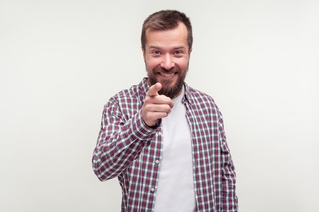 Ei, você Retrato de um homem barbudo alegre em camisa xadrez casual, apontando o dedo para a câmera e parecendo feliz e alegre, gesticulando que você é um incrível estúdio interno isolado no fundo branco