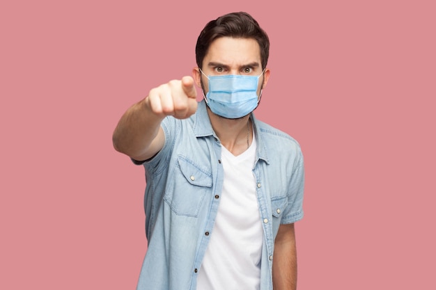 Ei você. Retrato de um homem sério com máscara médica cirúrgica em pé de camisa azul estilo casual apontando e olhando para a câmera com cara de bravo. tiro de estúdio interno, isolado no fundo rosa.
