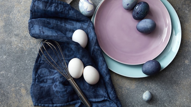Eier gemalt auf farbigen Tellern mit Schneebesen und Tuch auf einem Betontisch, flach