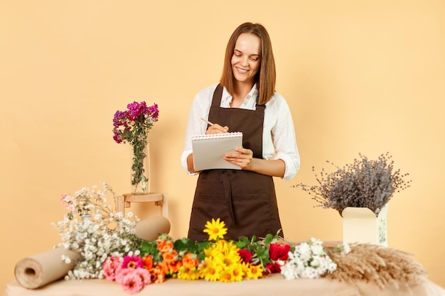 Eigentümer eines kleinen Blumengeschäfts, professioneller Blumenwerkstatt, Verkäuferin im Blumenladen, die eine Schreibtafel hält, schreibt und Notizen macht, Bestellung in einem Blumenladen gegen die beigefarbene Wand