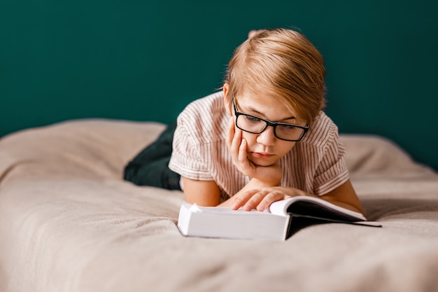 Ein 10-jähriger Junge mit Brille liegt auf dem Bett und liest ein großes Buch.