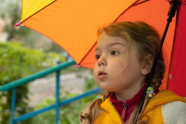 Ein 3-jähriges Mädchen versteckt sich im Regen unter einem farbigen Regenschirm