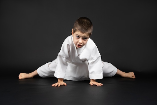 Ein 5-jähriger Junge übt Karate und streckt seine Beine auf schwarzem Hintergrund. Vorbereitung für das Training der Muskeldehnung