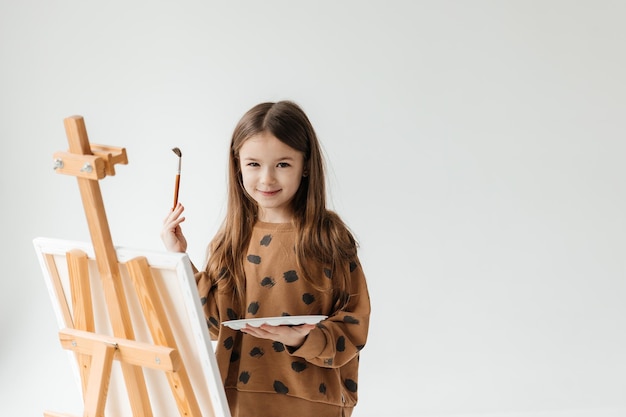 ein 6-jähriges Mädchen mit einem Pinsel in den Händen vor einem Staffel mit einer Leinwand auf einem isolierten weißen Hintergrund Studio Cyclorama