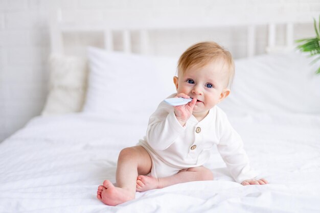 Ein 6 Monate alter blonder Junge nagt an einem Kamm auf einem weißen Bett in einem hellen Schlafzimmer in einem Baumwoll-Bodysuit, an dem ein kleines Kind zahnt