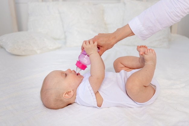 Ein 6 Monate altes Baby liegt auf einem weißen Bett und trinkt Milch aus einer Flasche, Babynahrungskonzept