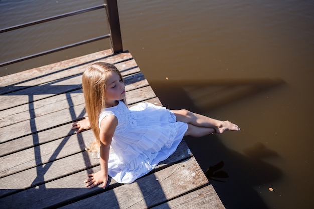 Ein 7-jähriges Mädchen mit langen blonden Haaren am See sitzt auf einer Clutch mit Beinen im Wasser. Sie spritzt ihre Füße in den See. Barfüßiges Mädchen in einem weißen Kleid mit langen Haaren.