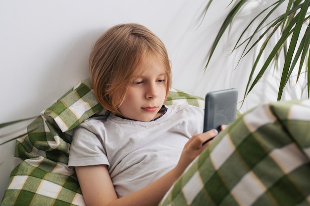 Ein 910-jähriger Junge mit langen Haaren liegt im Bett, vertieft in sein Mobiltelefon, erkundet die digitale Welt und spielt, was das Konzept von Entspannung und Nichtstun verkörpert