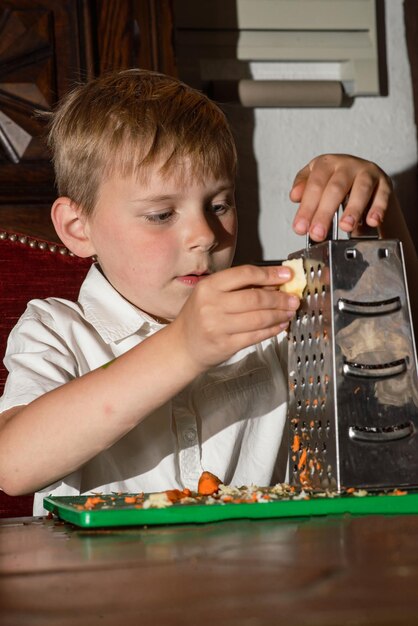 Ein achtjähriger Junge bereitet einen Salat zu, reibt ihn auf einer Reibe und schneidet ein echtes Foto