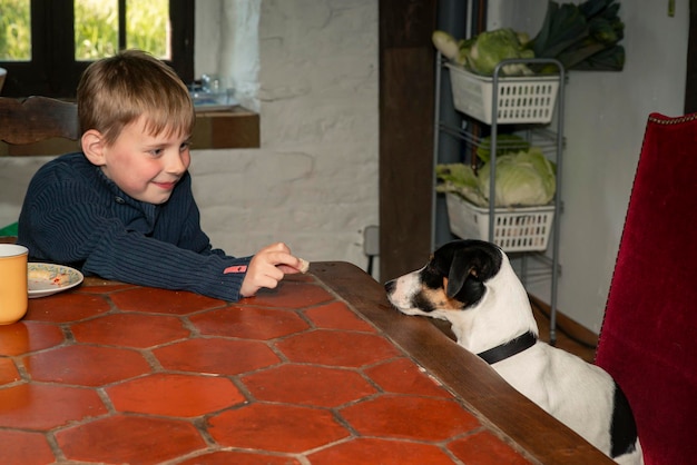 Ein achtjähriger Junge isst mit einem Hund an einem Tisch