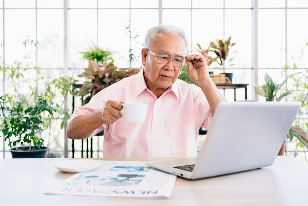 Ein älterer alter Mann im Ruhestand, der in rosafarbenem Freizeitstil gekleidet ist, benutzt einen Laptop, während er Kaffee trinkt
