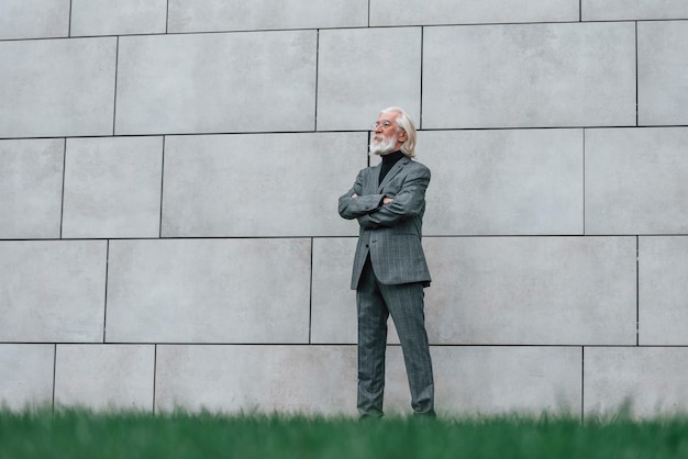 Ein älterer Geschäftsmann in formeller Kleidung mit grauem Haar und Bart steht im Freien an der Wand