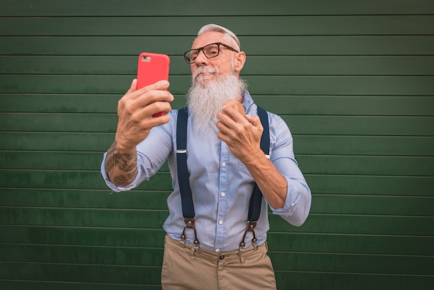 Ein älterer Mann älterer Mann in Hipster-Kleidung und Brille und ein langer weißer Bart kämmen seinen Bart und machen ein Foto