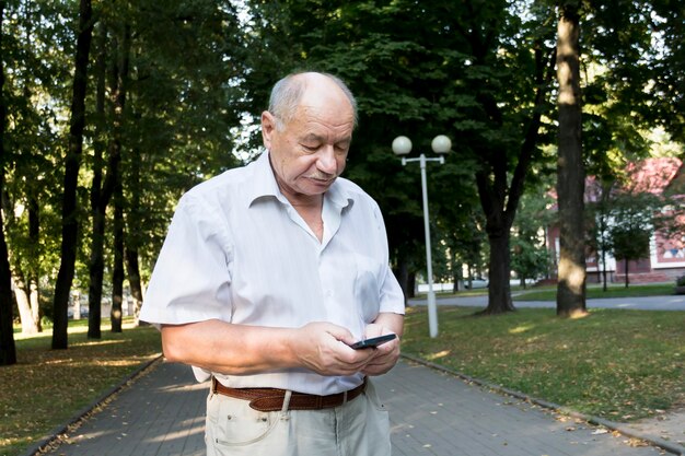 Ein älterer Mann geht im Sommer allein im Park Ein moderner Geschäftsmann im Ruhestand in weißem Hemd und Hose schreibt eine Nachricht und studiert Informationen auf einem Mobiltelefon