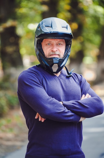 Ein älterer Mann im dunklen Helm im Freien. Freizeitkleidung. Gekreuzte Hände. Blauer Pullover. Unscharfer Hintergrund im Freien. Nahaufnahme.