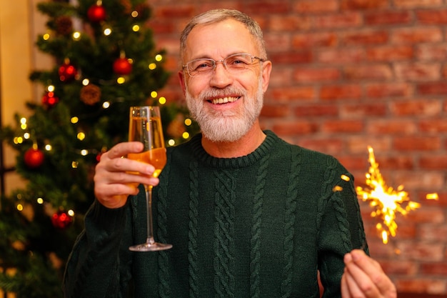 Ein älterer Mann in einem grünen Strickpullover, der neben dem Weihnachtsbaum einen Toast mit Champagner erhebt