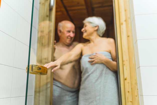 Ein älterer Mann und eine Frau im Ruhestand, die in Handtücher gewickelt sind und in die Spa-Sauna aus Holz gehen