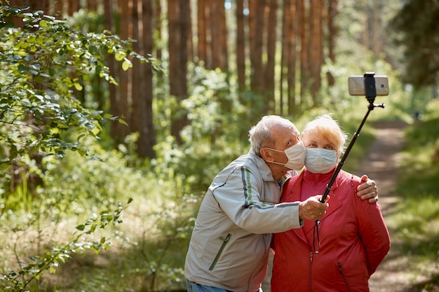 Ein älteres Ehepaar in Schutzmasken macht ein Selfie auf einem Smartphone und geht im Park spazieren