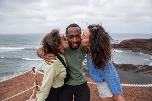 Ein Afrikaner erhält während eines Urlaubs einen Kuss von zwei kaukasischen Mädchen, eine glückliche gemischtrassige Gruppe hat Spaß.