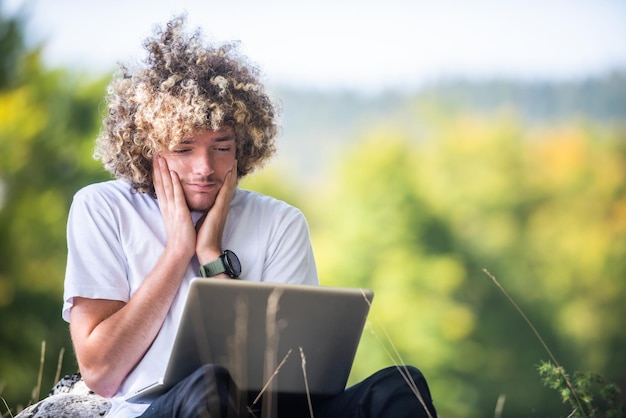 Ein Afro-Mann mit enttäuschtem Gesicht, der in der Natur sitzt und während einer Coronavirus-Pandemie einen Laptop für ein Online-Meeting benutzt