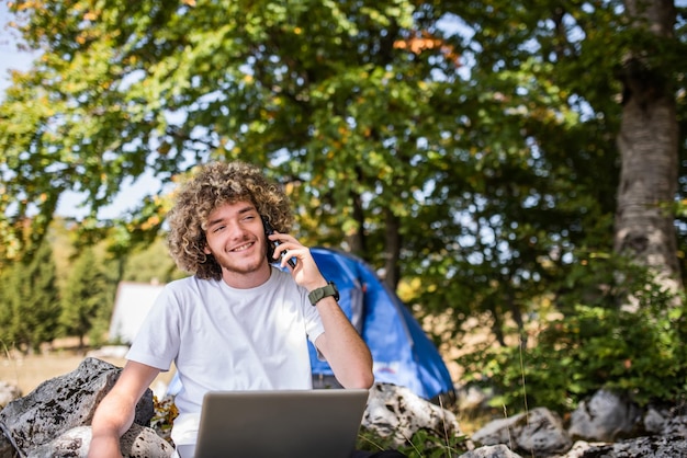 Ein Afro-Mann sitzt in der Natur und nutzt ein Smartphone und einen Laptop für ein Online-Meeting. Foto in hoher Qualität