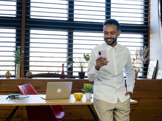 Ein afroamerikanischer Mann mit einem Laptop und einem Telefon in seinen Händen, der an einem Tisch im Büro sitzt, arbeitet online