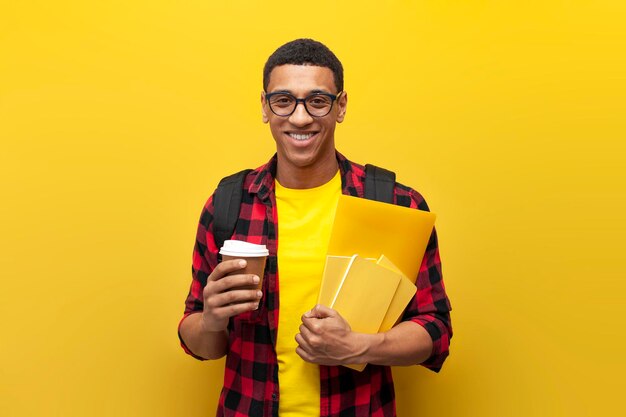 Foto ein afroamerikanischer student mit brille und rucksack hält kaffee und bücher auf gelbem hintergrund