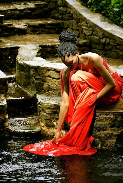 Ein afroamerikanisches Mädchen in einem roten Kleid sitzt auf einem Felsen in einem Park an einem Bach und wäscht ihr Kleid