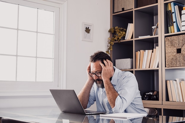 Ein alleiner unglücklicher und trauriger Mann zu Hause im Büro, der einen Laptop benutzt und ihn mit den Händen im Kopf betrachtet. Gestresster Mann in Schwierigkeiten bei der Lösung eines Problems