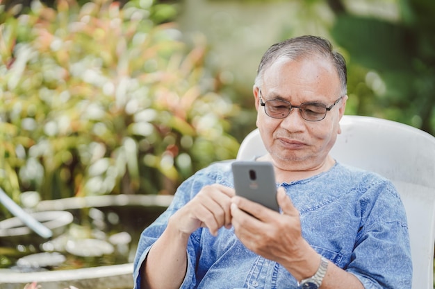 Ein alter Mann benutzt ein Smartphone auf einem Stuhl im Garten Soziales Netzwerkkonzept