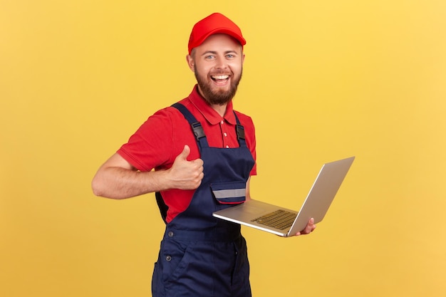 Ein Arbeiter mit blauen Overalls, der an einem Laptop mit Daumen nach oben arbeitet, mag einen neuen Onlinedienst