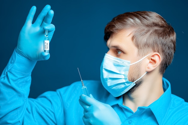 Ein Arzt in einer medizinischen Maske und Handschuhen, gekleidet in eine blaue Uniform, hält eine Injektionsspritze und eine Ampulle mit einem Impfstoff gegen Coronavirus vor sich